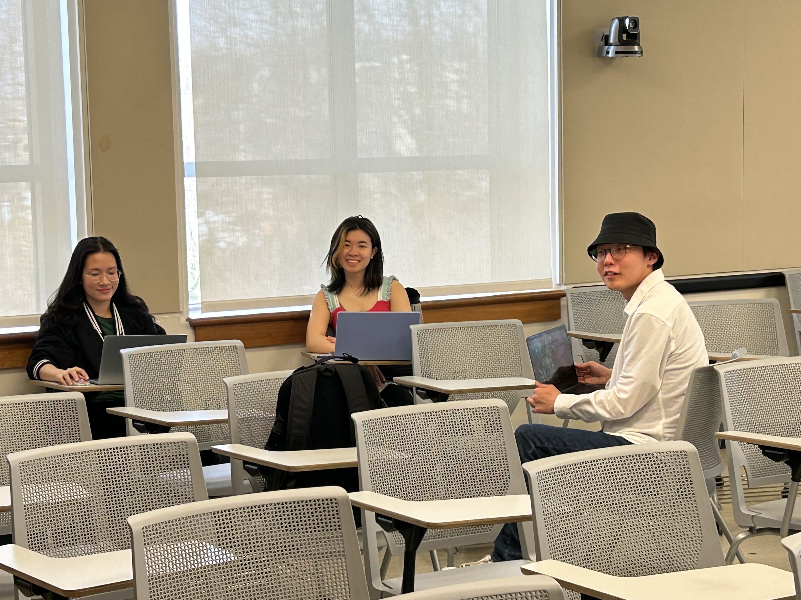 Two students sitting and looking at laptop screens, and one student standing and looking at the camera in a classroom full of rolling chairs.