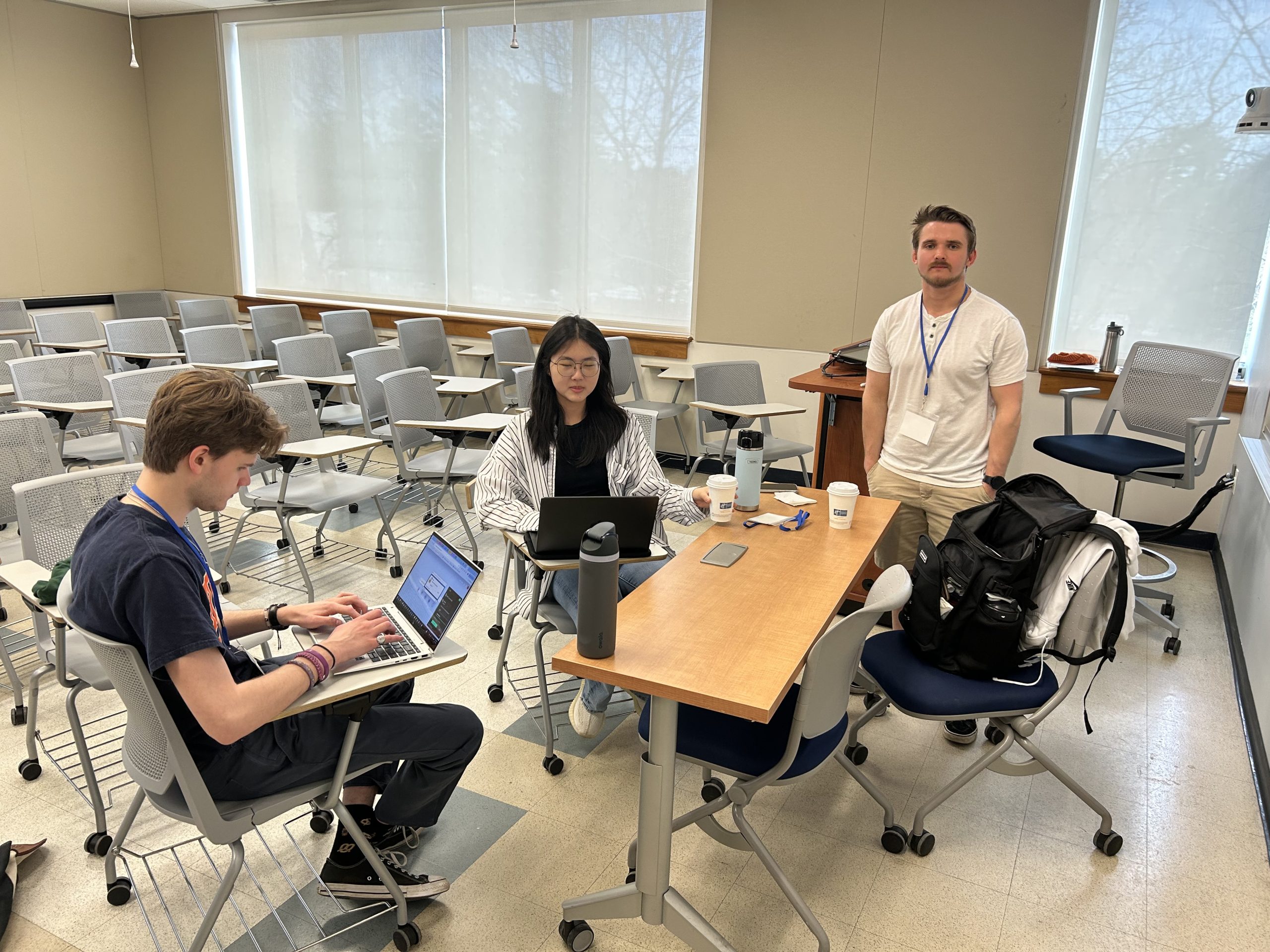 Two students sitting and looking at laptop screens, and one student standing and looking at the camera in a classroom full of rolling chairs.