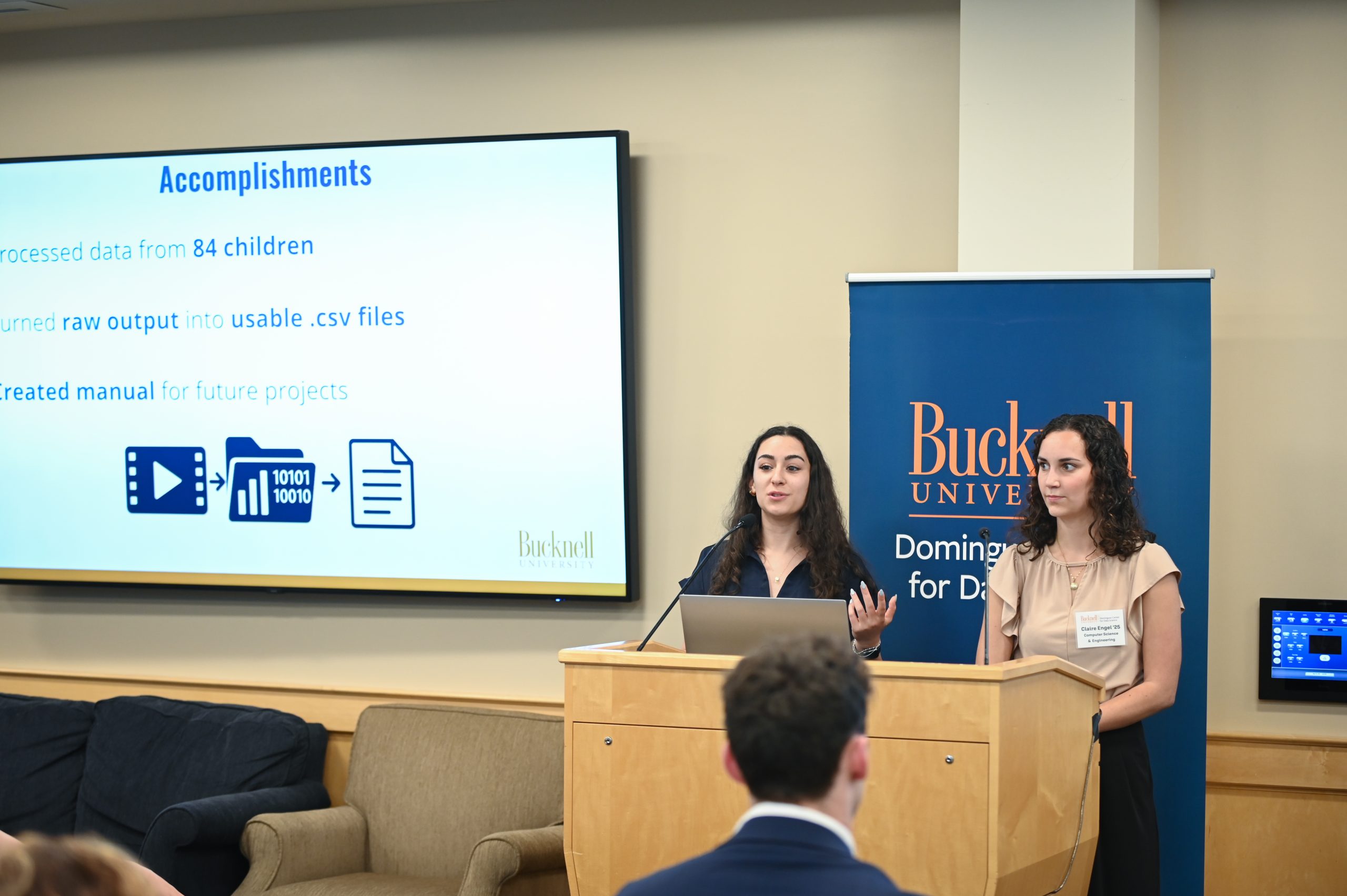 Claire Engel and Gwen Radecki standing behind a podium next to a screen that reads, "Acomplishments."