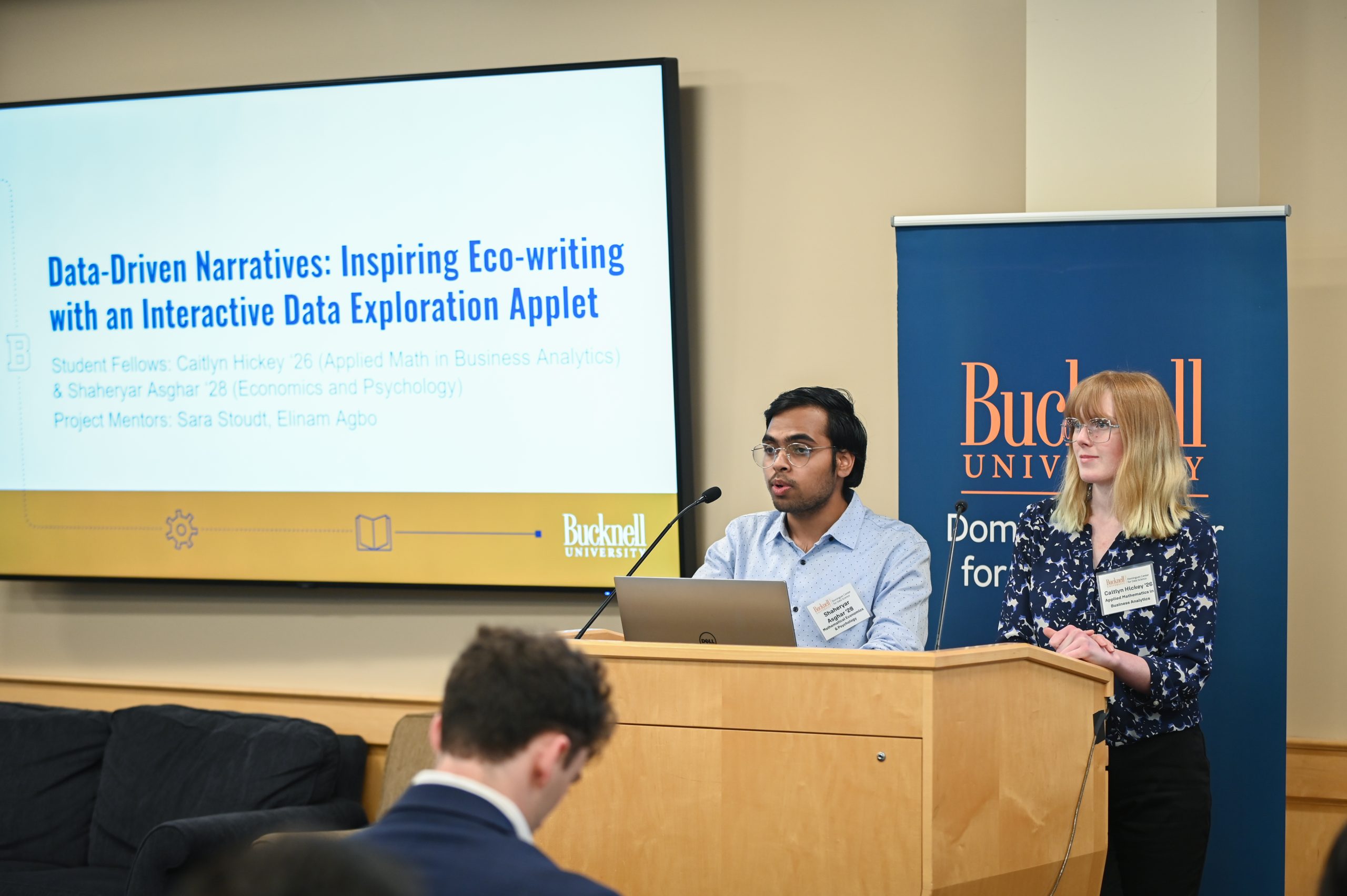 Claire Engel and Gwen Radecki standing behind a podium next to a screen that reads, "Acomplishments."