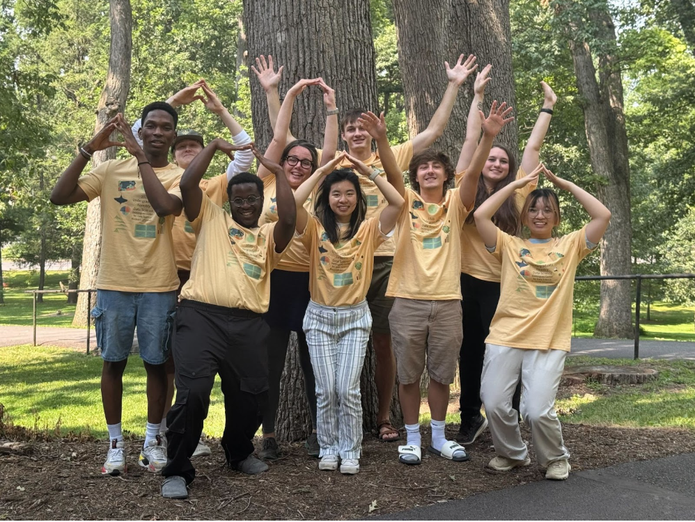 A group photo with all of the students and mentors who worked on the Bucknell Undergraduate Forestry Data Science Research Program wearing matching yellow shifts, standing in front of trees.