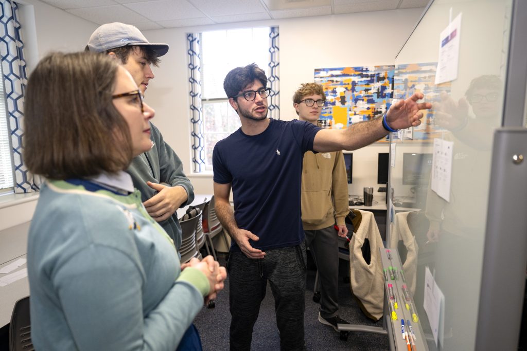 A student pointing at a whiteboard and explaining the paper there to a small group.