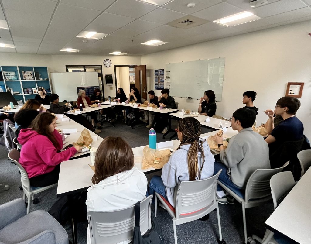 A large circle of tables, with students eating lunch and listening to Laurie Nelson speak at the front.