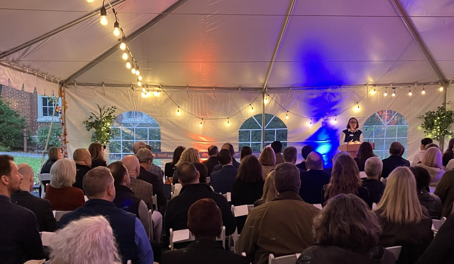 Kelly McConville standing behind a podium and speaking to a large crowd of guests lit by handing lights in a tent.